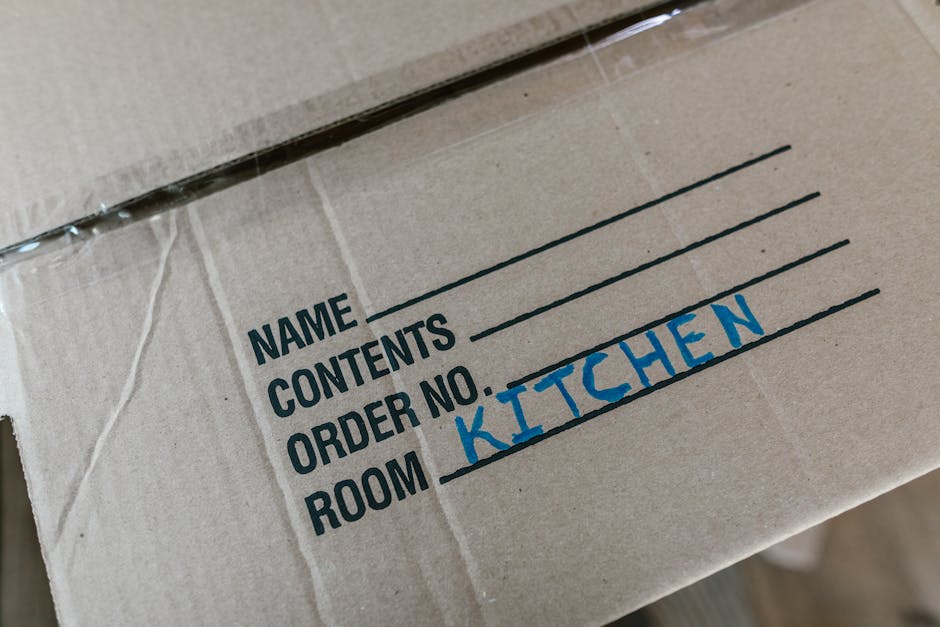 Close-up view of a cardboard moving box labeled with printed sections for 'Name,' 'Contents,' 'Order No.,' and 'Room,' with the handwritten word 'KITCHEN' in blue ink filling the 'Room' field. The box appears to be inside a property, situated on a flat surface with other packing materials nearby, indicating the process of packing and packing materials used for home relocation. The packaging is made of brown corrugated cardboard, and the handwriting suggests an organized approach to furniture transport or packing and moving. The background is slightly out of focus, emphasizing the label with natural lighting highlighting the box's surface, reinforcing the context of professional removals and house moving services provided by Removal Companies Ilford, supporting an SEO relevant description for house removals and relocation.