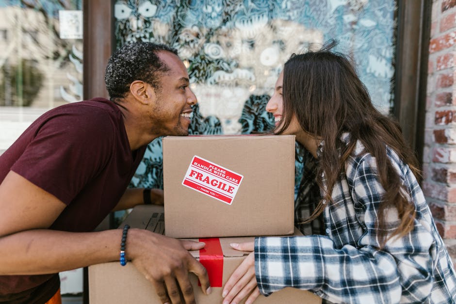 A man and a woman are engaged in a home relocation process outdoors, standing close together and smiling while exchanging a cardboard box. The man, with short curly hair and wearing a maroon T-shirt, is holding a large moving box with a red 'FRAGILE' sticker, indicating careful handling. The woman, with long dark hair and wearing a checkered shirt, is receiving the box with both hands. They are positioned in front of a glass door or window, reflecting trees or outdoor scenery, suggesting they are either preparing to load or unload the items for a house move. Additional packed boxes, some wrapped in plastic or cardboard, are partially visible, along with a cart or trolley used to assist in carrying heavy items. The scene is well-lit with natural daylight, emphasizing the collaborative and organized nature of the packing and moving process, which is part of the comprehensive home removal services offered by Removal Companies Ilford, supporting efficient furniture transport and packing logistics.
