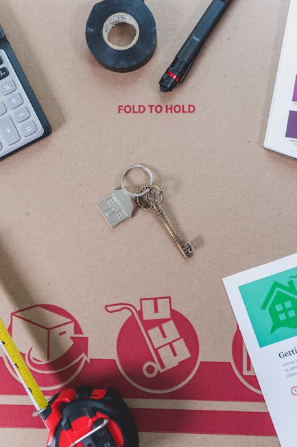 An overhead view of a cardboard moving box used in home relocation, with various packing and moving tools arranged on its surface. Visible items include a roll of black and blue packing tape, a black marker, a keyring with a small house-shaped charm and a vintage-style brass key, a yellow tape measure, and a calculator. Part of a printed packing checklist or instructions can be seen at the edge of the box, along with a partial view of a document with colorful graphics. The box appears to be inside a property, serving as a workspace for packing or preparing items for a house move, reflecting the process of furniture transport and packing logistics. The scene is well-lit, emphasizing the tools and materials necessary for a professional removal service, as might be handled by companies such as Removal Companies Ilford during a detailed house removals process.