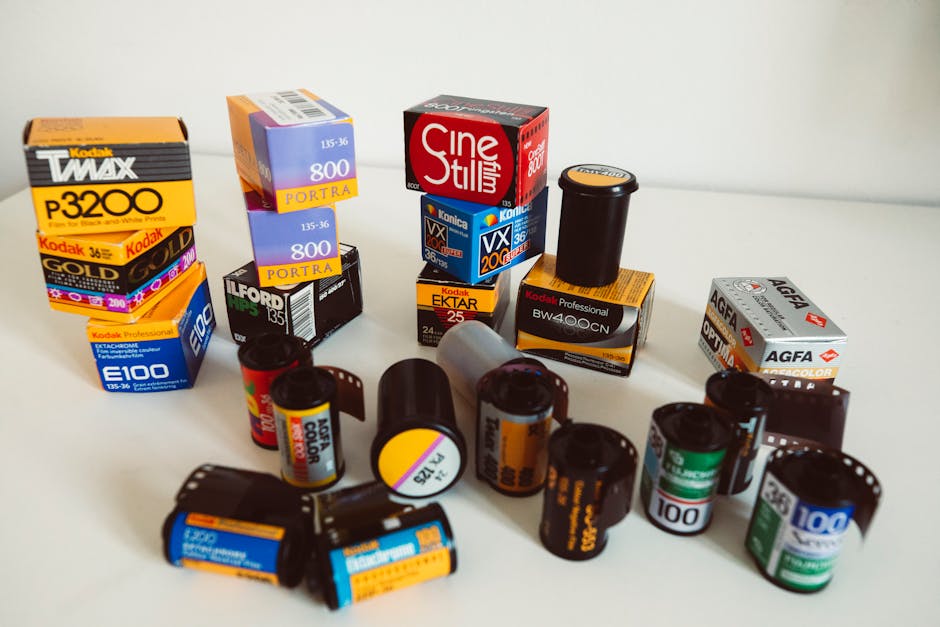 An array of photographic film rolls and film canisters are arranged on a plain, light-colored surface inside a property, likely in preparation for a home relocation or packing process. The collection includes various types of film, such as 35mm, 120mm, and instant film canisters, with some in cardboard boxes and others loose. Notable brands visible include Kodak, Agfa, and Ilford, with labels indicating different film types like color negative, black and white, and professional-grade film, many with bright packaging and distinctive coloured labels. The film rolls are scattered in a semi-organised manner, with some standing upright and others lying horizontally. An opened cardboard box of Kodak Professional Ektar film, along with other boxes and individual rolls, suggest a packaging and handling process occurring during a move. Although the scene primarily focuses on film storage, it subtly supports the theme of packing, transportation, and furniture or belongings handling during a move, consistent with removals services provided by [COMPANY_NAME]. The setting is well-lit with an even light, emphasizing the details on each film container, and the background is plain, keeping the focus on the objects involved in the packing and moving activity.