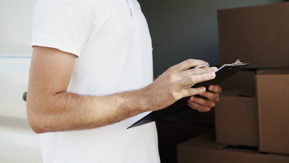 Close-up view of a person wearing a white t-shirt holding a black clipboard with a pen, standing next to a stack of packed brown cardboard boxes and wrapped furniture in an interior setting. The boxes are stacked on a ground surface, and some are covered with plastic wrapping, indicating they are prepared for moving or relocation. In the background, there is a dark wall and additional packing materials visible, suggesting a home or storage environment. The scene captures the packing and moving process typical of house relocations, with a focus on inventory or checklist management by Removal Companies Ilford in preparation for furniture transport and home relocation services, as seen on their website page for booking cancellations and rescheduling.