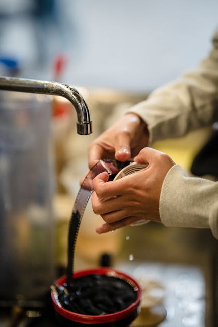 Inside a residential property, a person wearing a beige long-sleeved shirt is holding a flexible black garden hose and connecting it to a metal sink tap, likely in preparation for moving or cleaning d