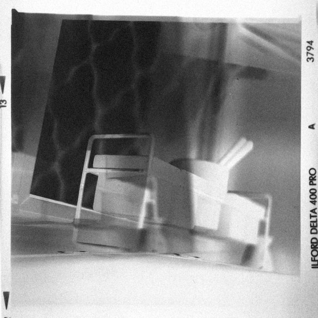 Black and white photographic slide showing a home relocation scene, featuring a small metal trolley with a handle and wheels positioned on a tiled floor near a doorway. The trolley holds several cardboard boxes, some wrapped in plastic or fabric covers, with additional boxes placed nearby on the floor. One of the boxes contains long objects, possibly packing materials or tools. The background reveals part of a wall and a door frame, indicating an indoor setting. The image captures the process of packing and preparing household items for transport, consistent with furniture transport and packing activities undertaken by removals companies like Removal Companies Ilford, as part of professional house removals or shifting services. The scene emphasizes logistics involved in moving household contents, highlighting packing materials, boxes, and trolley equipment used during the loading process.