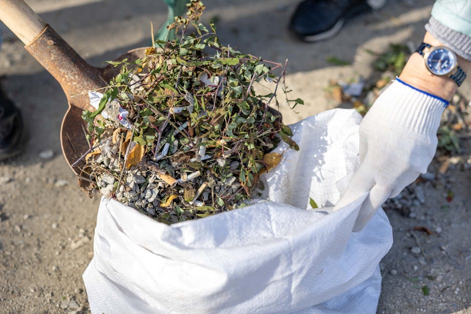 A person wearing a white glove and blue wristwatch is holding a large white sack filled with garden debris including small branches, leaves, and cigarette butts, while standing outdoors on a dirt surface. Nearby, a metal shovel with a wooden handle leans into the sack, which is partially open to reveal the contents. The background shows a paved area with a pair of black shoes and another person's foot visible, suggesting a collaborative effort in a house removal or property clearance process. The scene captures the loading or disposal phase of a home relocation project, emphasizing proper handling of garden waste in accordance with local waste rules, as indicated by the relevant web page about Ilford removals and bulky waste public regulations.