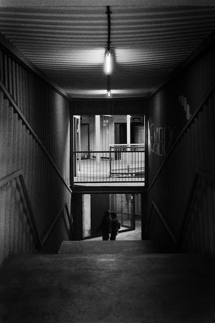 Black and white photographic slide showing a home relocation scene, featuring a small metal trolley with a handle and wheels positioned on a tiled floor near a doorway. The trolley holds several cardboard boxes, some wrapped in plastic or fabric covers, with additional boxes placed nearby on the floor. One of the boxes contains long objects, possibly packing materials or tools. The background reveals part of a wall and a door frame, indicating an indoor setting. The image captures the process of packing and preparing household items for transport, consistent with furniture transport and packing activities undertaken by removals companies like Removal Companies Ilford, as part of professional house removals or shifting services. The scene emphasizes logistics involved in moving household contents, highlighting packing materials, boxes, and trolley equipment used during the loading process.