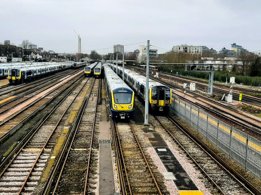 A view of multiple modern passenger trains parked on parallel railway tracks in a railway yard, with some trains featuring yellow front panels and others in grey and white colour schemes. The tracks are laid with wood and metal sleepers, and a metal fence runs alongside the tracks on the right side of the image. Overhead electrical lines supported by metal poles stretch across the scene, with a cityscape of buildings, including residential and commercial structures, visible in the background under an overcast sky. This scene illustrates a typical railway environment suitable for a home relocation service illustrating aspects of transport logistics. Removal Companies Ilford may use such images to convey their expertise in moving furniture and household items via train or to showcase their knowledge of transportation infrastructure for relocations between areas like Ilford station and local parks, such as Valentines Park.