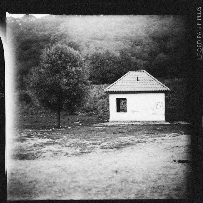 A black-and-white photograph of a small, single-story house with a gabled roof made of tiles, situated on a dirt or gravel ground. The house has a single window on the front facade, which appears to b