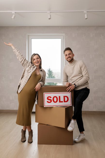 A cheerful couple stands inside a bright, empty room with light-colored wallpaper and a large window behind them, demonstrating their recent home sale. The woman, wearing a brown dress and checkered b