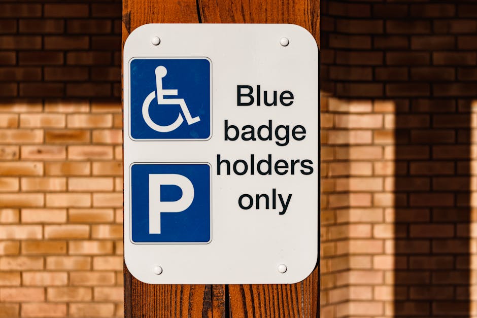 A rectangular white parking permit sign mounted on a wooden post outdoors, with a blue accessibility symbol at the top indicating space reserved for wheelchair users. Below, there is a blue parking symbol with a white 'P' and text that reads 'Blue badge holders only' in black letters. The sign is positioned against a background of a brick wall, with sunlight casting a shadow on the wall and part of the sign. This signage is relevant to house removals and furniture transport services, such as those provided by Removal Companies Ilford, which often require parking considerations for home relocation activities. The sign indicates designated parking restrictions in residential or commercial areas, reflecting the importance of planning for proper permissions and parking permits involved in the moving process, especially when space is limited or parking rules are strict.