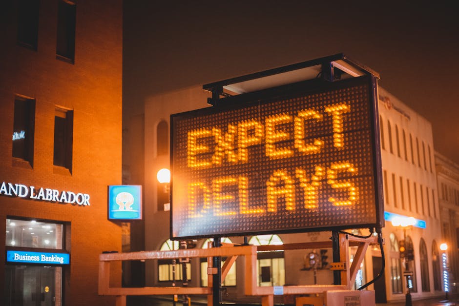 A large electronic road sign displays the message 'EXPECT DELAYS' in bright orange LED lights, positioned on a portable stand on a city street at night. The background includes illuminated buildings with commercial signage, such as a bank and a business center, indicating a busy urban environment. The sign is placed near the pavement, with visible metal framework and wiring supporting it. The scene reflects a context of traffic management or disruption, relevant to house removals and moving logistics, where delays in transportation might affect scheduled home relocation or furniture transport services. The street lighting and the glow of the LEDs emphasize the urban setting, with other smaller lights emanating from shop windows and nearby street lamps.
