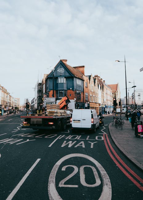 A white flatbed truck operated by Removal Companies Ilford is parked in the middle of a city street, actively engaged in a home relocation or furniture transport process. The truck's bed is loaded with wooden and cardboard boxes, wrapped furniture, and packing materials, which are secured and ready for moving. The truck is positioned behind a white van, which is assisting in the loading or unloading activities at a busy urban intersection. Surrounding the scene are multi-storey commercial and residential buildings, with some showing brick facades and a mix of modern and traditional architecture. Several pedestrians are walking along the sidewalk, some near bicycles and parked scooters, while traffic lights and street signage indicate a central area within Ilford. Overhead, a partly cloudy sky provides natural light, illuminating the scene, which captures the logistical aspects of a professional removals service, including packing, loading, and transportation as part of a house or office move. The environment reflects an active urban setting where removals specialists from Removal Companies Ilford are managing the logistics of home or furniture transport efficiently.