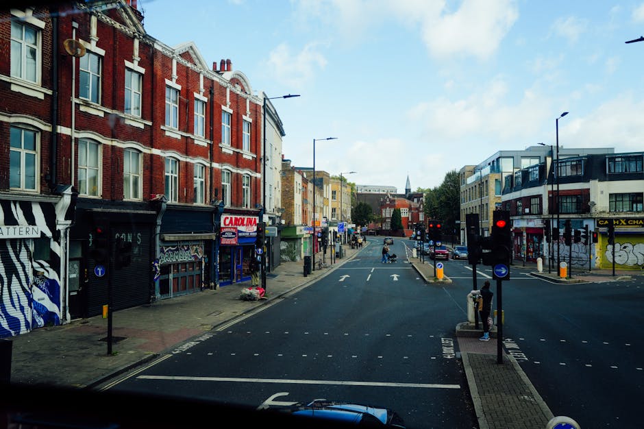 The image shows a street scene in a commercial area with a mix of brick and modern buildings, some with shopfronts and signage, on both sides of the road. The street is relatively busy with a few pedestrians, and several cars and vans are visible, including a small commercial vehicle parked on the pavement near the curb. Traffic lights are present at the intersection, currently displaying red signals. On the right side, a person is standing near the traffic light, and another is walking along the pavement. The scene includes street furniture such as bollards and lamp posts. The lighting indicates daytime with partly cloudy skies, providing natural light that illuminates the street uniformly. This setting aligns with the context of house removals or furniture transport processes, possibly on a route advice guide from Seven Kings to Ilford High Road, coordinated by Removal Companies Ilford for efficient moving logistics and packing activities.