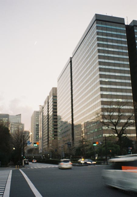 A city street scene featuring a modern, multi-storey office building with reflective glass windows and horizontal paneling, situated at a busy intersection during daytime. In the foreground, blurred moving vehicles are visible on the road, with a few cars stationary at traffic lights. Leafless trees line the pavement, and the sky above is partly cloudy with a faint contrail. The urban environment includes other tall office buildings in the background, suggesting a commercial district. This scene exemplifies typical city infrastructure that Removal Companies Ilford might navigate during house or office relocation services, with the high-rise building representing a common destination or origin point for furniture transport and packing-related activities within their local removal routes from Seven Kings to Ilford High Road.