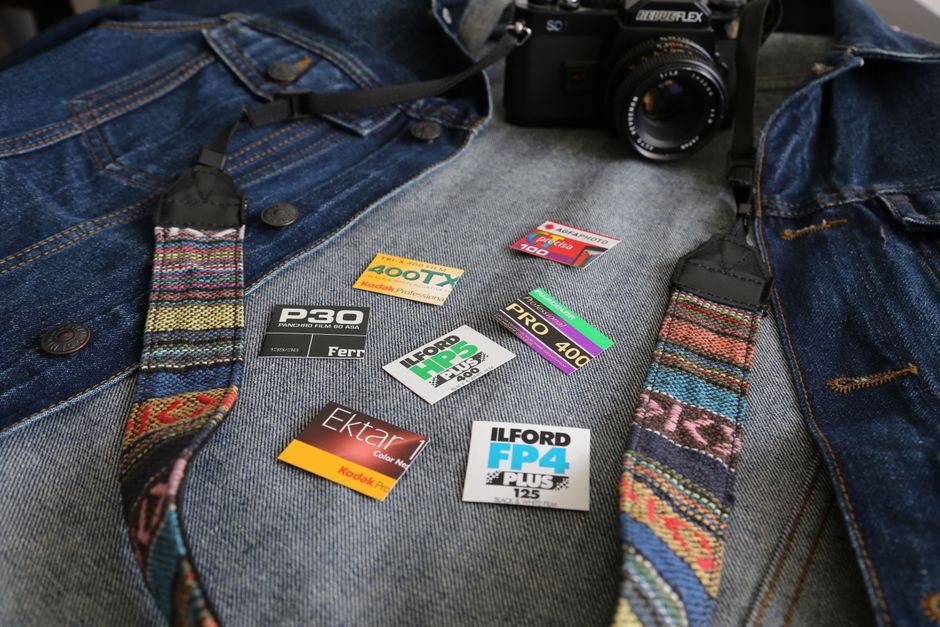 A flat lay photograph displays various photographic film and memory cards arranged on a grey fabric surface. The items include a black camera positioned at the top center, with its strap hanging down and partially obscured by the cards. Several SD cards and compact memory cards with colourful labels are spread out below and around the camera, featuring brand names such as Kodak and Ilford. The SD cards and memory cards are of different sizes and colours, including red, yellow, black, and purple. The scene appears to be indoors, with soft lighting illuminating the surface, which resembles the interior of a household or workspace suited for photography tasks. This arrangement of equipment and media cards demonstrates a focus on photography and media storage, relevant to the home relocation context, as businesses like Removal Companies Ilford often assist with packing and transporting such personal or professional equipment during moves. The composition emphasizes careful organisation of photographic accessories, suitable for detailed packing and moving considerations related to specialist equipment.